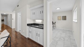 A spacious kitchen with white cabinets and a black countertop, a dining area with a bar stool, and a hallway with wooden flooring.
