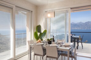 A dining area with a table set for four, large windows with blinds, a potted plant, and a view of the sea and mountains.