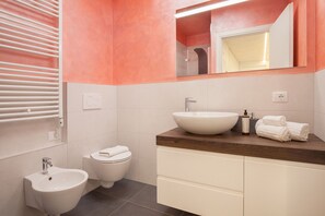 A modern bathroom with a white sink, a bidet, and a toilet. There is a wooden vanity with a mirror above the sink.