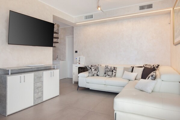 Una sala de estar moderna con un televisor de pantalla plana montado en la pared, un sofá blanco con cojines estampados, un mueble blanco con cajones y una cafetera sobre una encimera blanca.