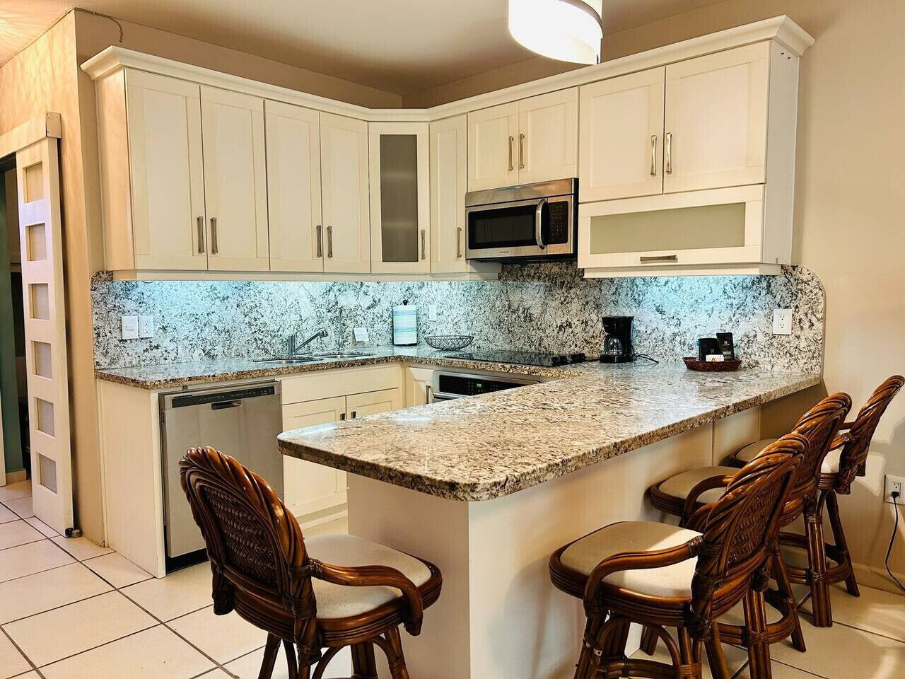 A kitchen with white cabinets, a granite countertop, a microwave, and a coffee maker. There are three wooden chairs around the island.