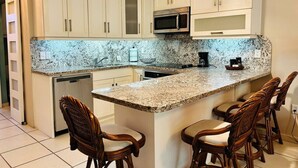A kitchen with white cabinets, a granite countertop, a microwave, and a coffee maker. There are three wooden chairs around the island.