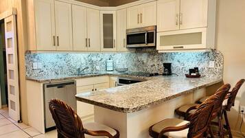 A kitchen with white cabinets, a granite countertop, a microwave, and a coffee maker. There are three wooden chairs around the island.