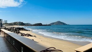 A coastal view with a sandy beach, rocky shoreline, and a distant mountain.