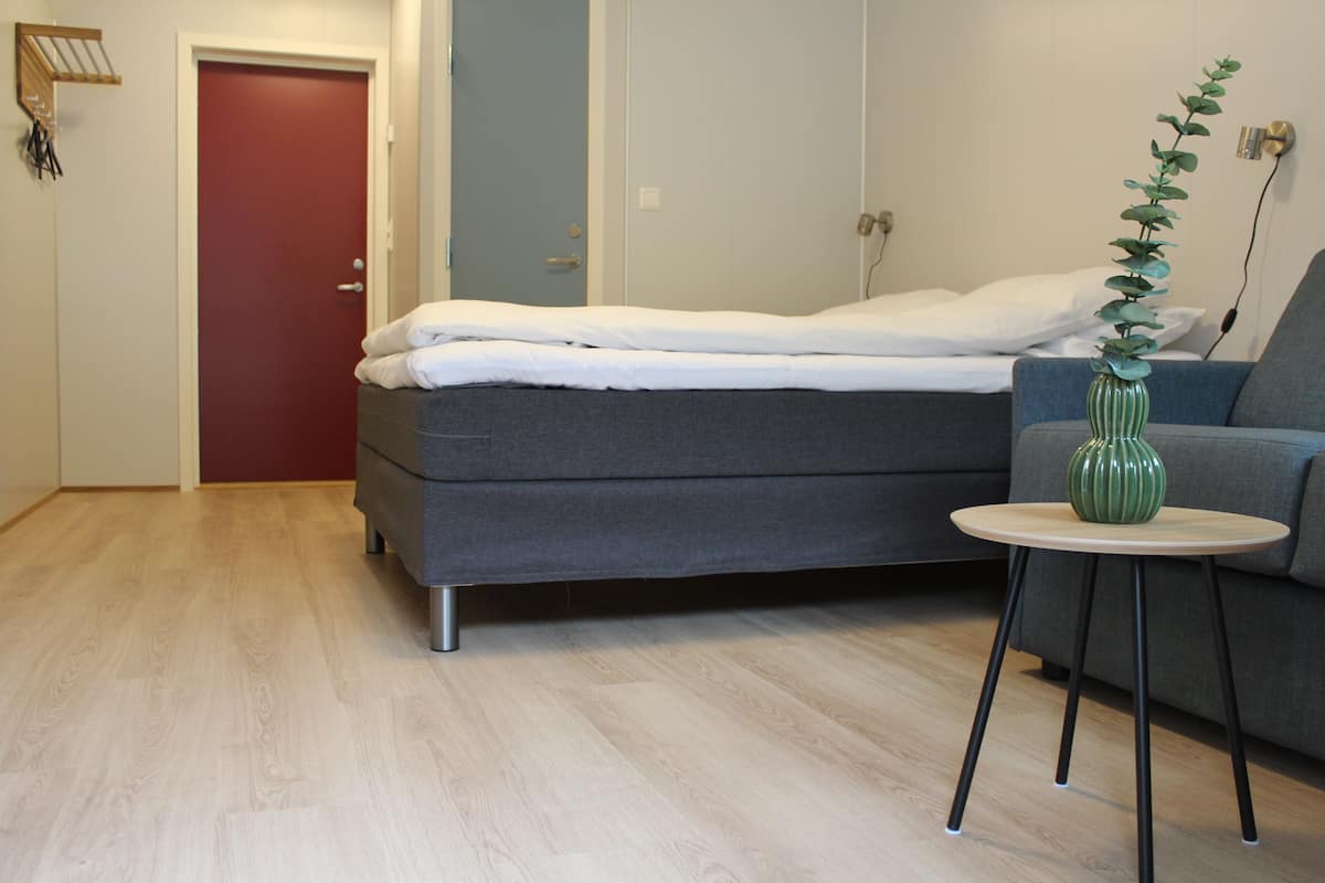 Quarto com cama, sofá cinza, mesa redonda pequena com um vaso verde e porta vermelha.