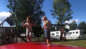 Two children jumping on a red trampoline in a backyard with a wooden play structure, trees, and a white fence. - Utvika Gård & Camping (Hole)