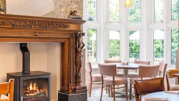 A room with a fireplace, a dining table with chairs, and large windows with a view of greenery.