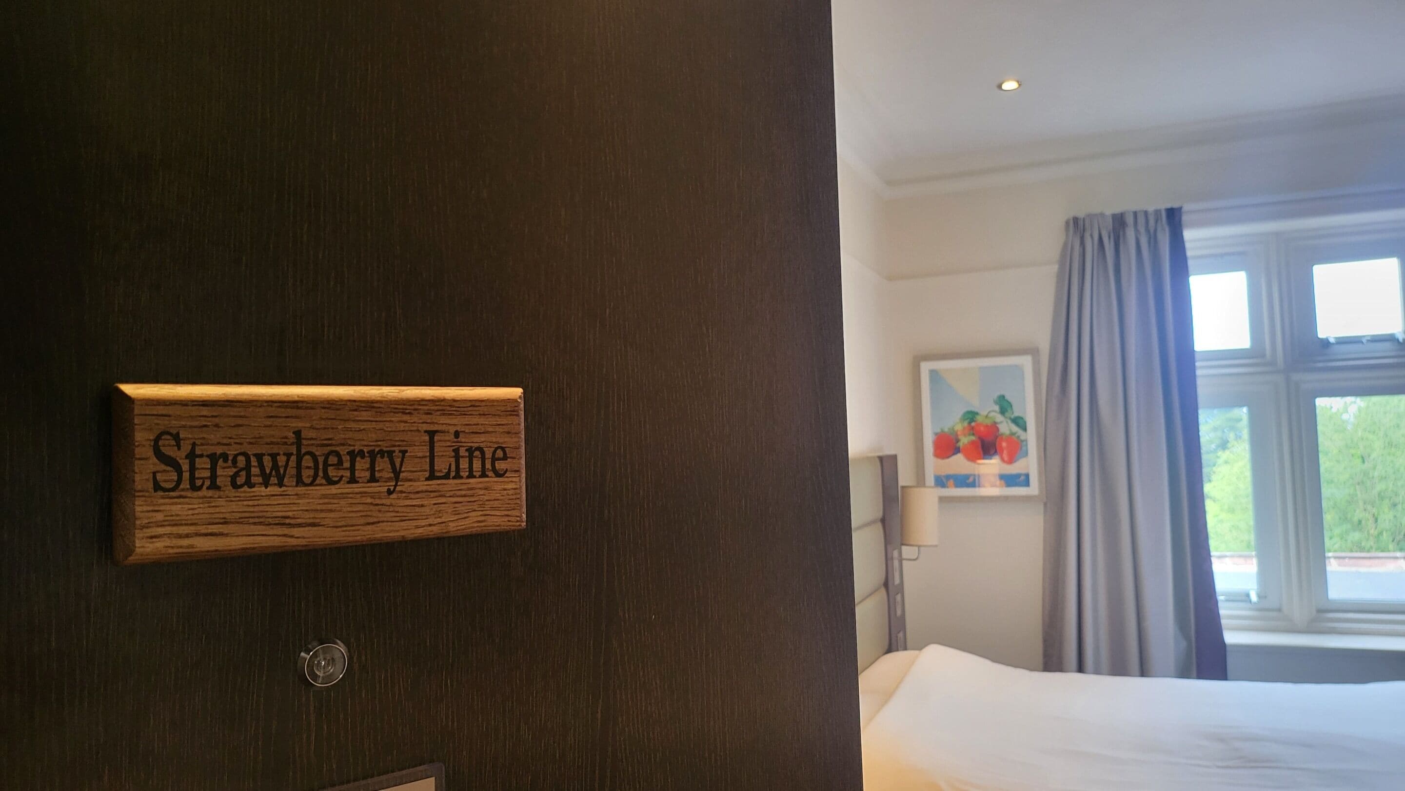 A hotel room with a wooden door labeled "Strawberry Line," a bed, a window with curtains, and a painting of strawberries.