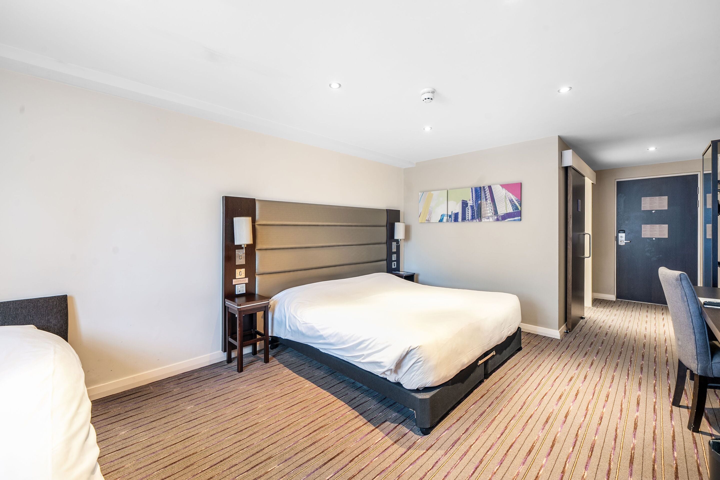 Ein Hotelzimmer mit einem großen Bett, einem Schreibtisch mit Stuhl, einem kleinen Tisch und einem Bild an der Wand.