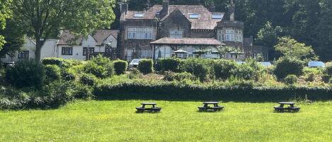 Ein großes, historisches Gebäude mit zahlreichen Fenstern und einer markanten Dachlinie, umgeben von üppigem Grün und Picknicktischen auf einer Wiese.