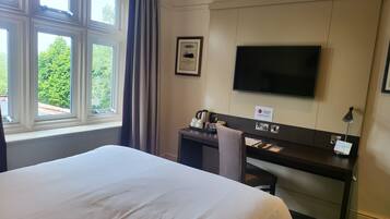 Habitación de hotel con cama, escritorio con televisor de pantalla plana, un cuadro en la pared y ventana con vista a árboles.