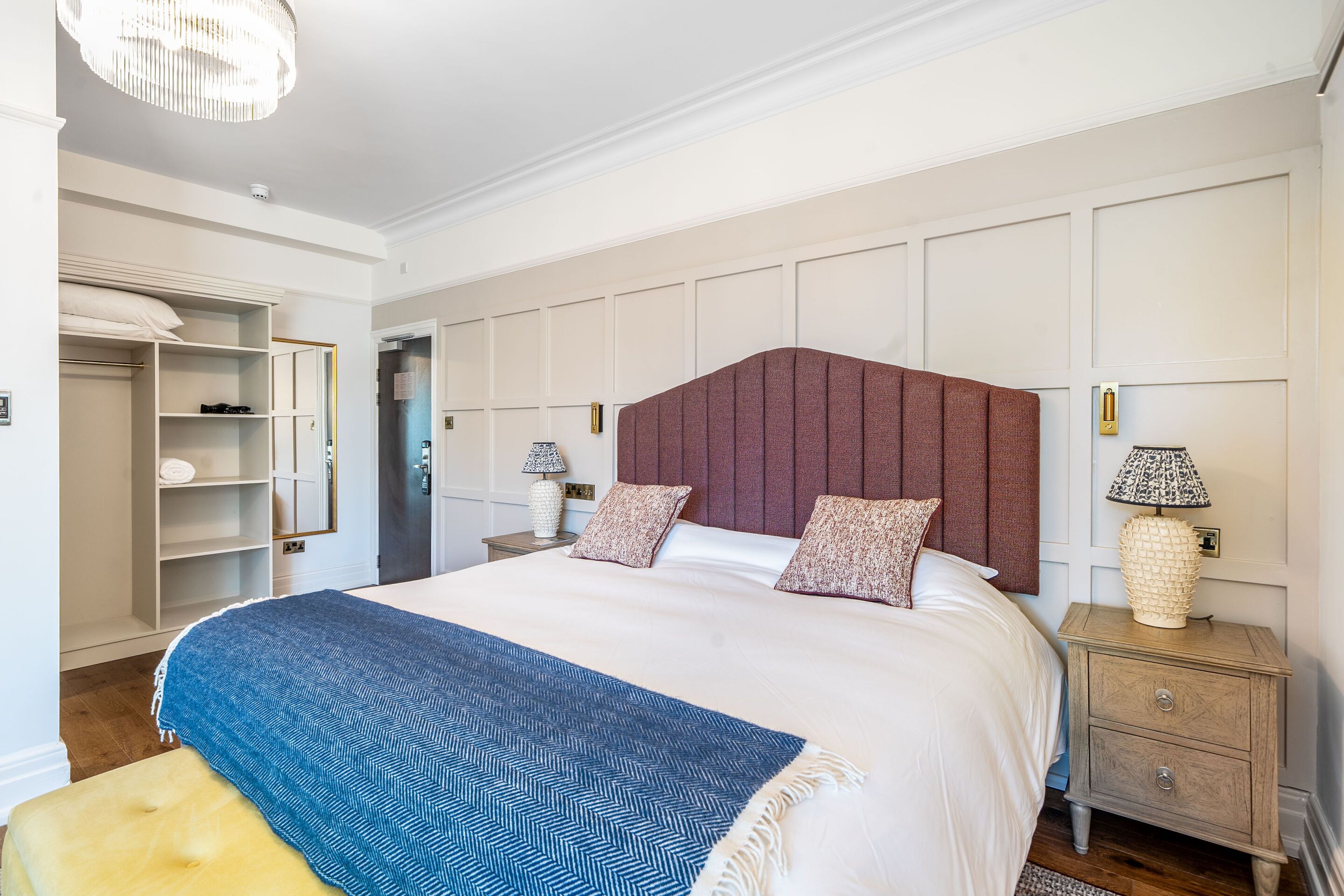 A neatly arranged bedroom with a large bed, a bedside table with a lamp, a wardrobe, and a door leading to another room.