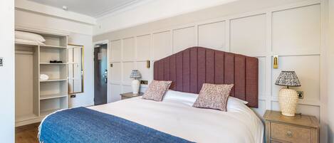 A neatly arranged bedroom with a large bed, a bedside table with a lamp, a wardrobe, and a door leading to another room.
