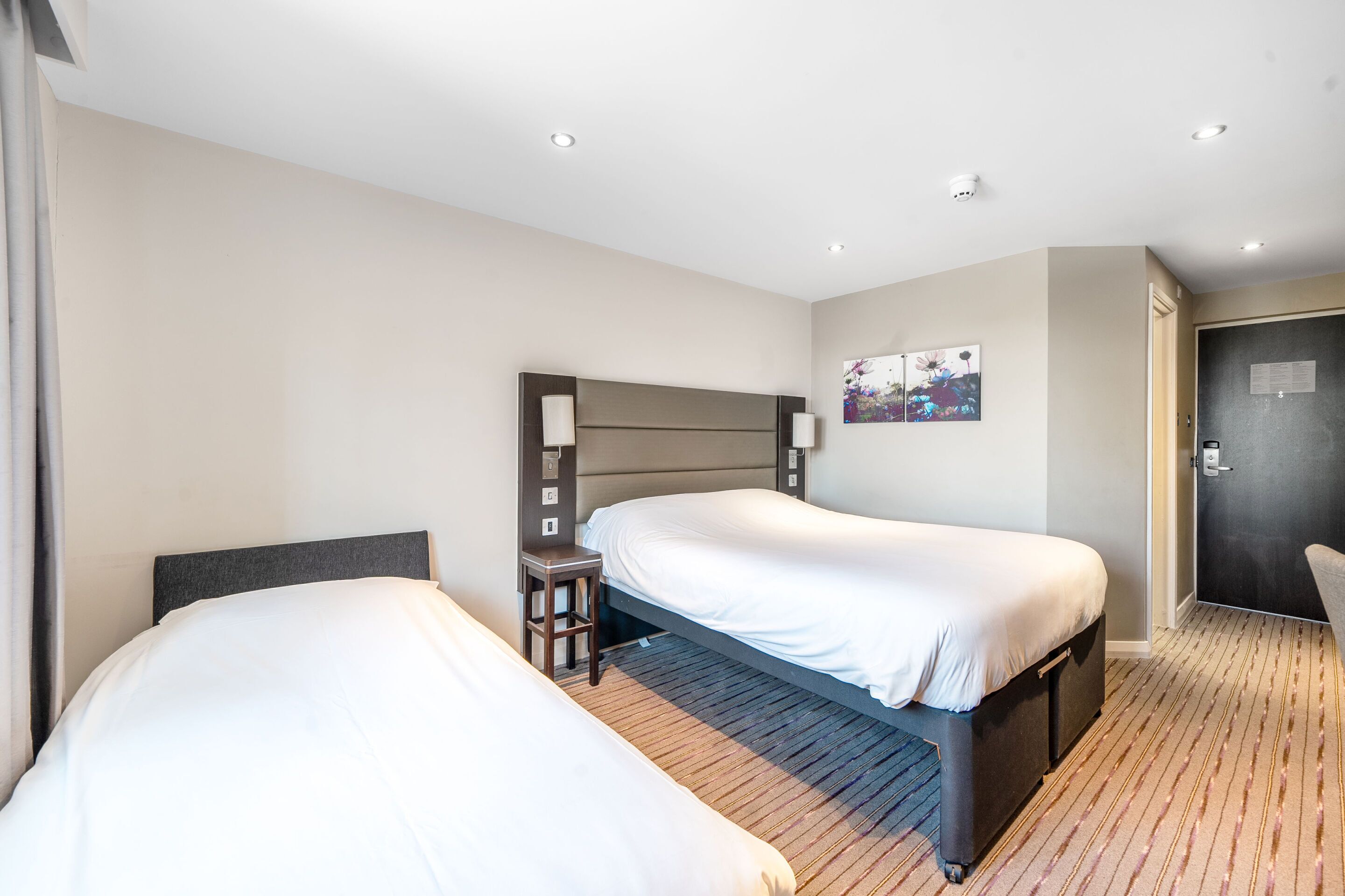 A hotel room with two single beds, a small table, a chair, and a painting on the wall.