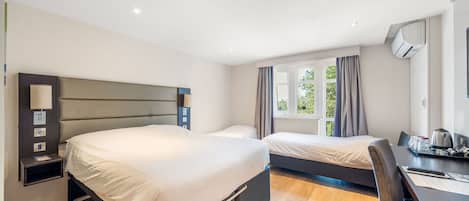 Quarto de hotel com uma cama grande, uma cama menor, uma escrivaninha com cadeira e uma janela com cortinas.