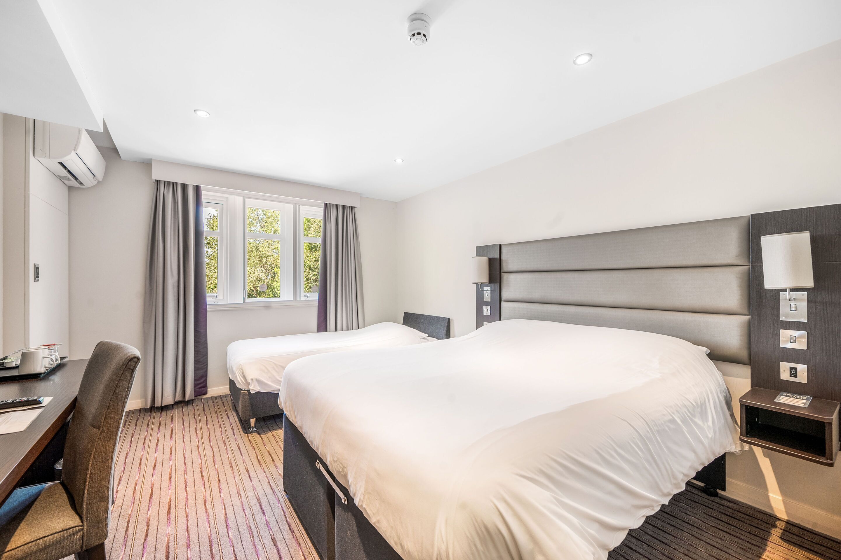 Habitación de hotel con dos camas, un escritorio, una silla, una mesita de noche con lámpara y una ventana con cortinas.