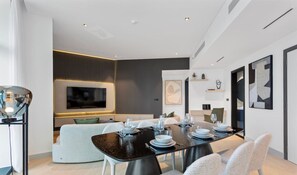 A modern dining area with a black table set for four, white chairs, and a large flat-screen TV mounted on the wall.