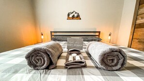 Una cama bien hecha con una colcha a cuadros, dos toallas enrolladas y un cartel que indica las comodidades de la habitación.