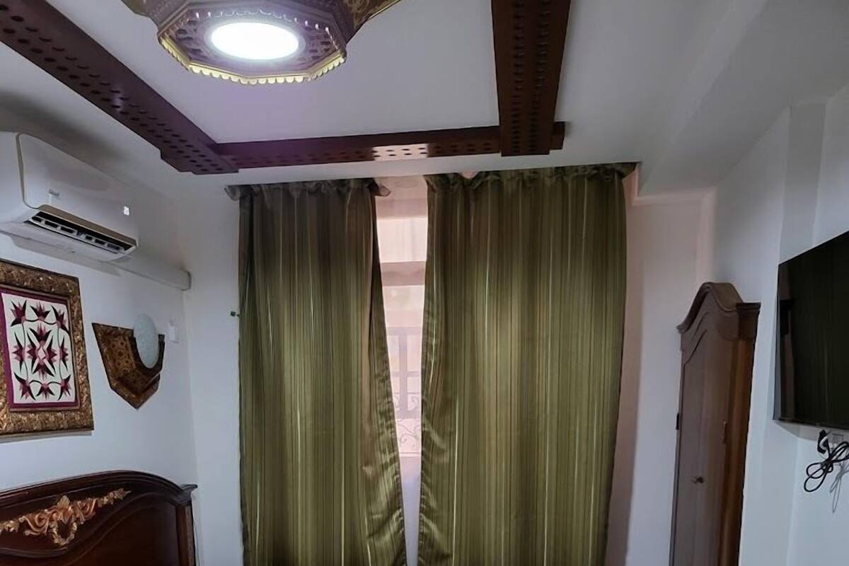A hotel room with a bed, a wooden nightstand, a television, and a window with green curtains.