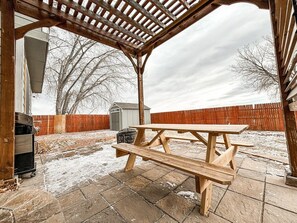 Una mesa de picnic de madera bajo una pérgola en un patio trasero con un cobertizo y una valla de madera al fondo.
