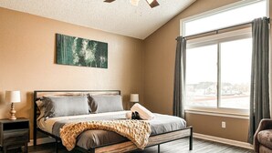Une chambre à coucher avec un lit, des tables de chevet, un ventilateur de plafond, une fenêtre avec des rideaux et un tableau au mur.