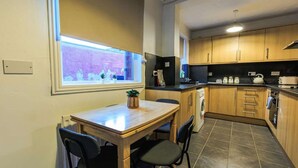A compact kitchen with wooden cabinets, a dining table with chairs, a washing machine, and a window with a view of a purple wall.