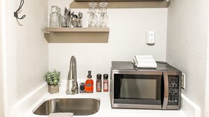 Cocina compacta con un lavamanos, un microondas y estantes superiores con vajilla y cristalería.