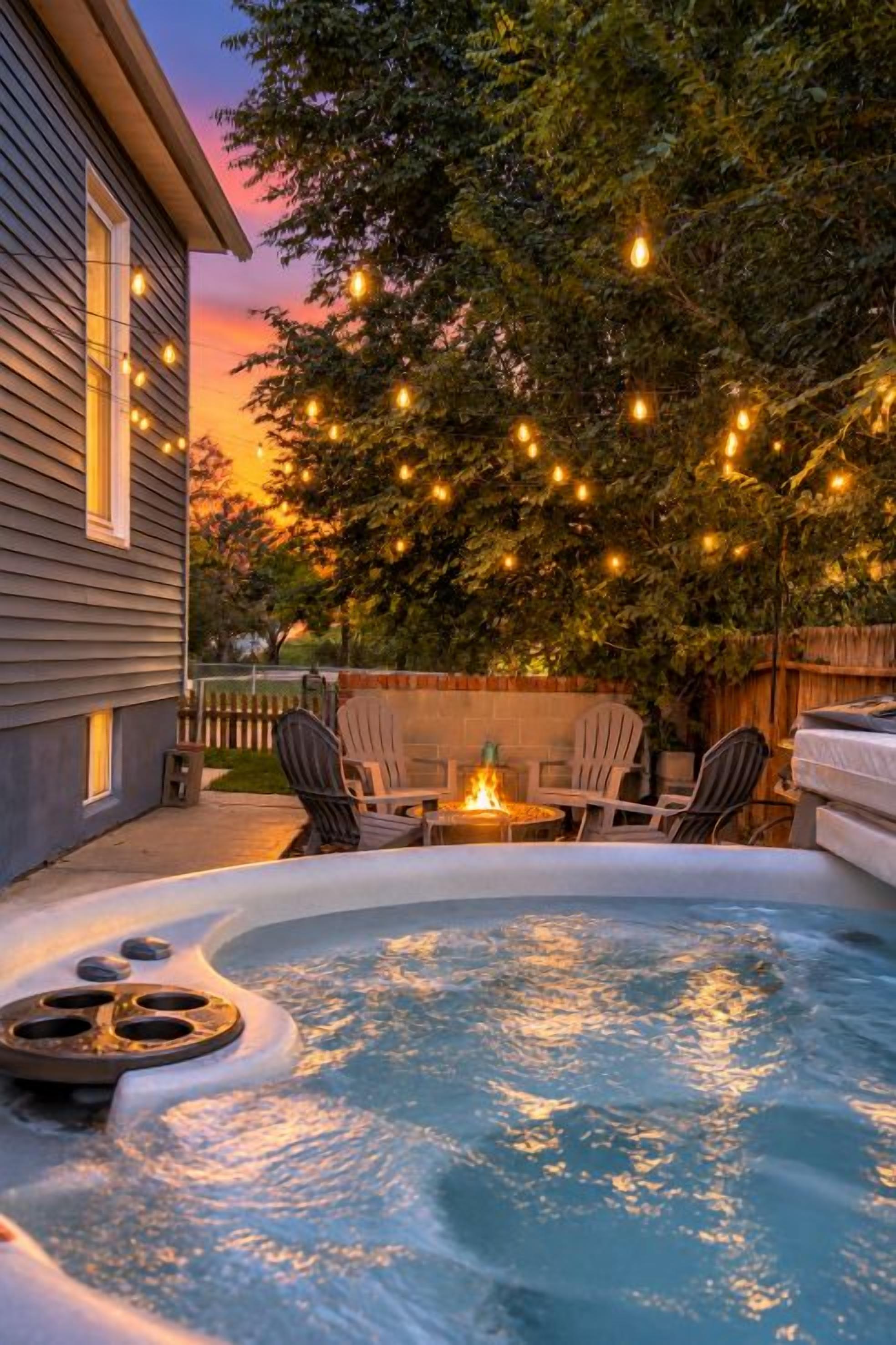 Een jacuzzi met vuurplaats, omringd door tuinmeubilair en sfeerverlichting, met op de achtergrond bomen en een huis.