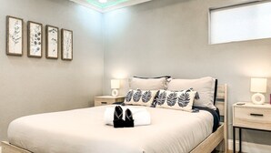 Ein ordentlich bezogenes Bett mit weißer Decke und schwarzen Hausschuhen, zwei Nachttische mit Lampen sowie vier gerahmte Pflanzenbilder an der Wand.