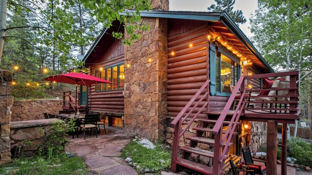 Una cabaña de troncos con fachada de piedra, una terraza de madera, mobiliario exterior y una escalera que lleva a un porche.