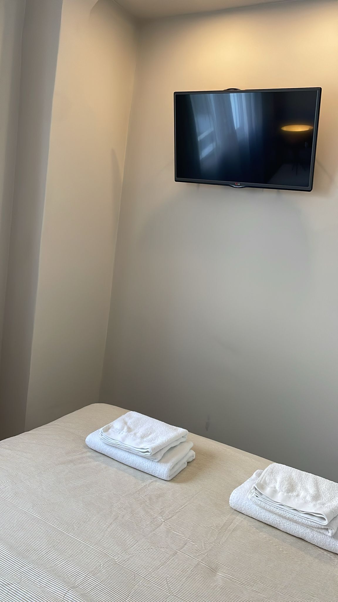 A hotel room with a bed, a flat-screen TV mounted on the wall, and two folded white towels on the bed.