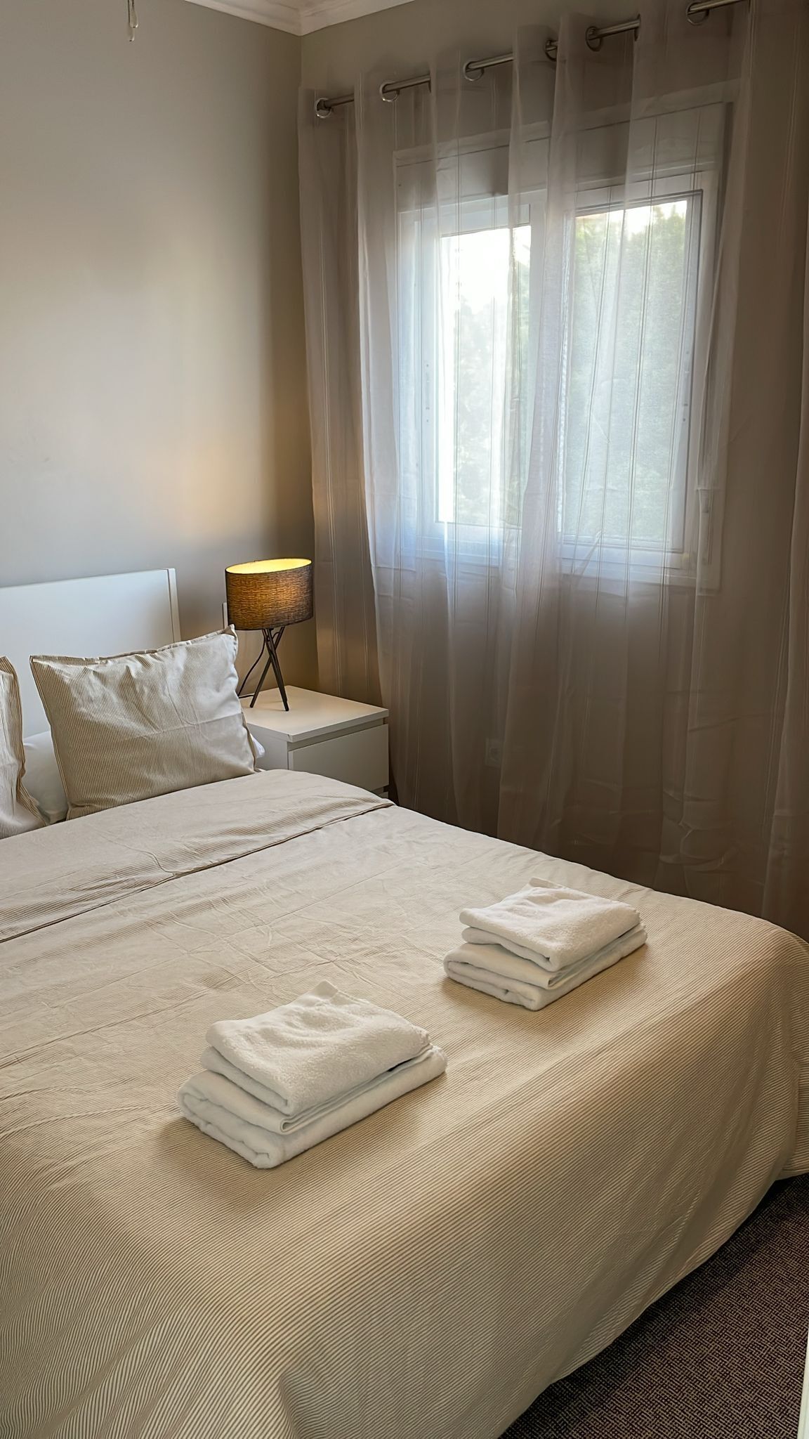 Een net opgemaakt bed met witte lakens en handdoeken, een bedlamp en dunne gordijnen voor een raam.