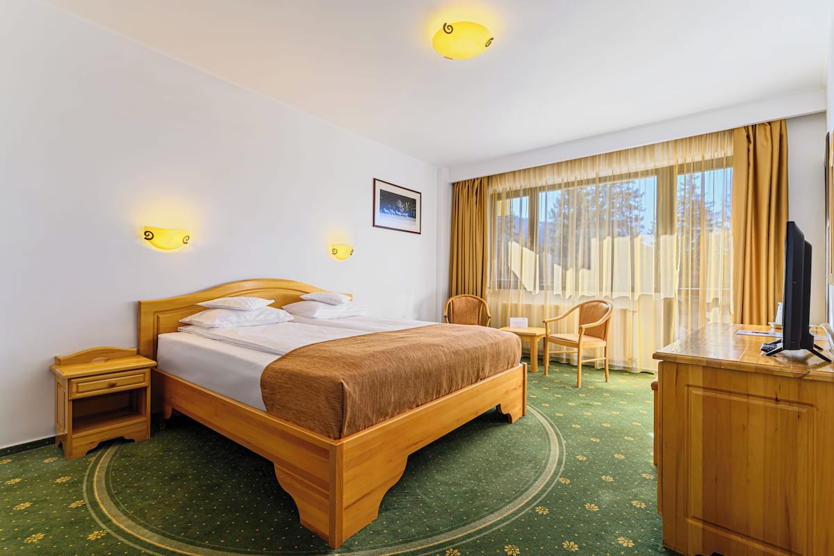 Ein Hotelzimmer mit einem hölzernen Bett, einem Schreibtisch mit Fernseher, einem Sessel und einem Fenster mit Vorhängen.