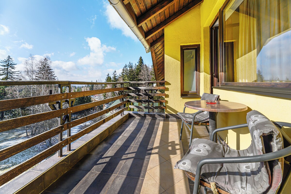 A balcony with a wooden railing, a table with chairs, and a view of trees and a river.