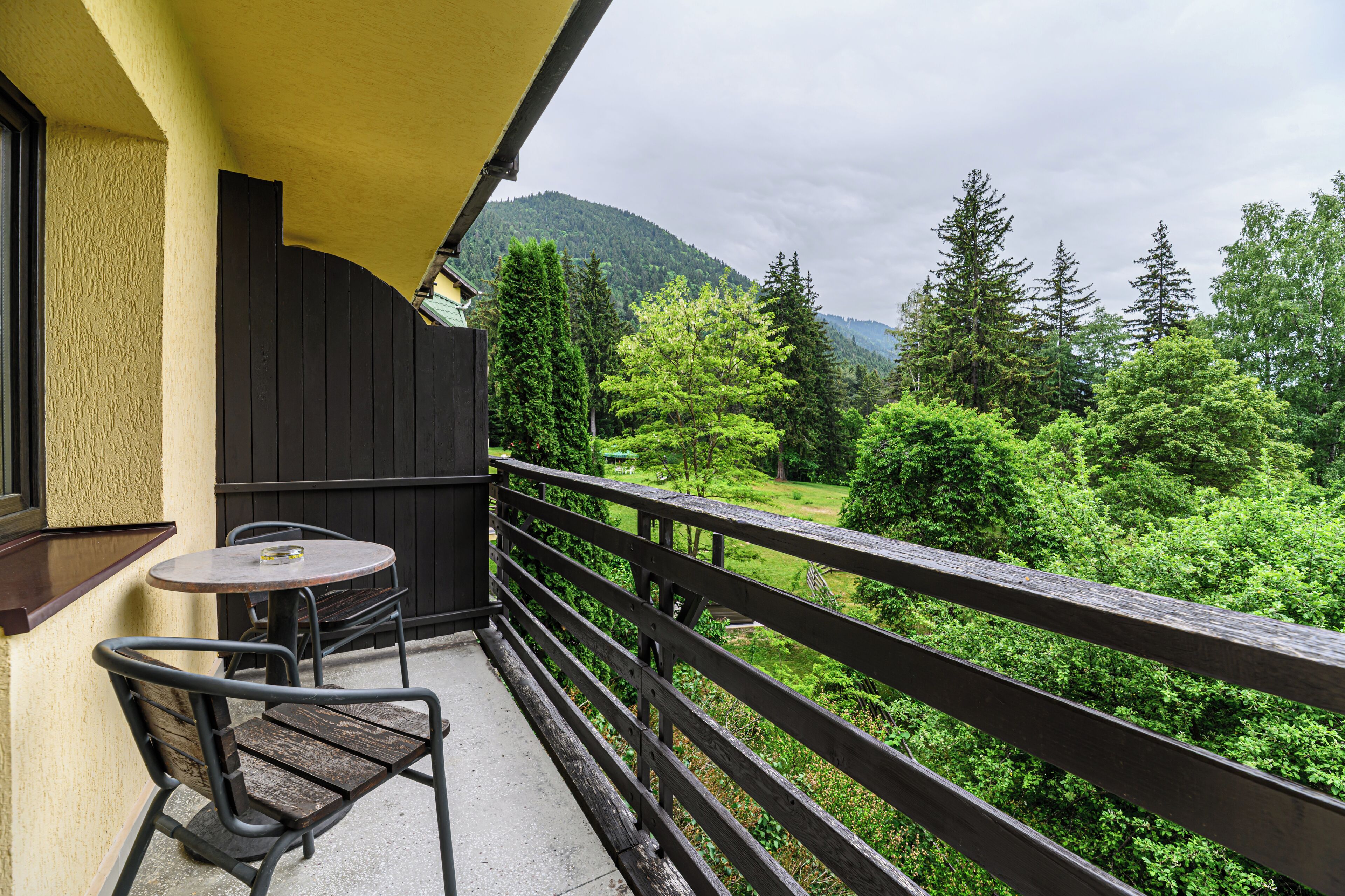 A balcony with a table and chair, overlooking a forested area with mountains in the background.