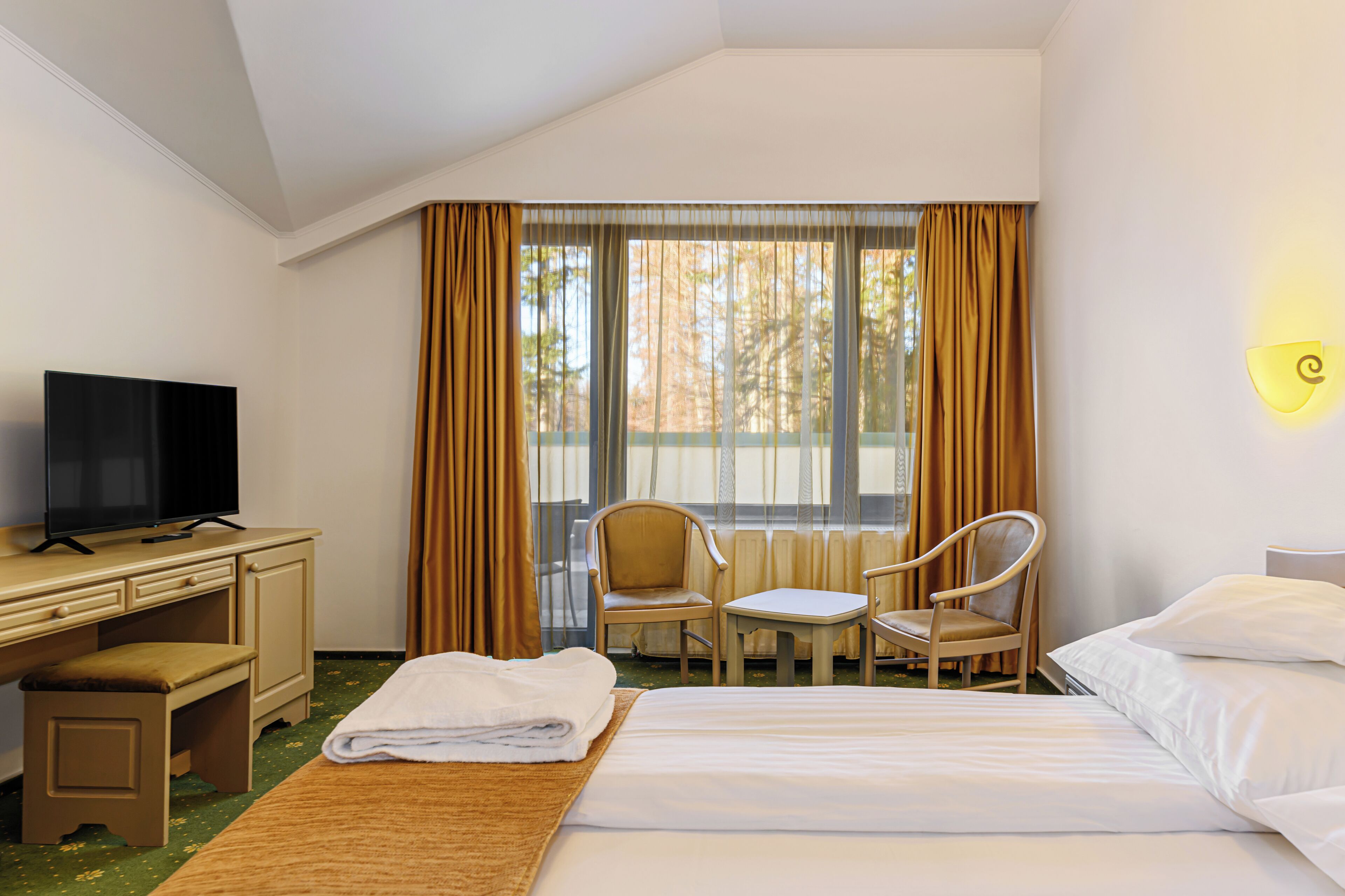 Habitación de hotel con cama, escritorio con televisor, dos sillas y ventana con cortinas.