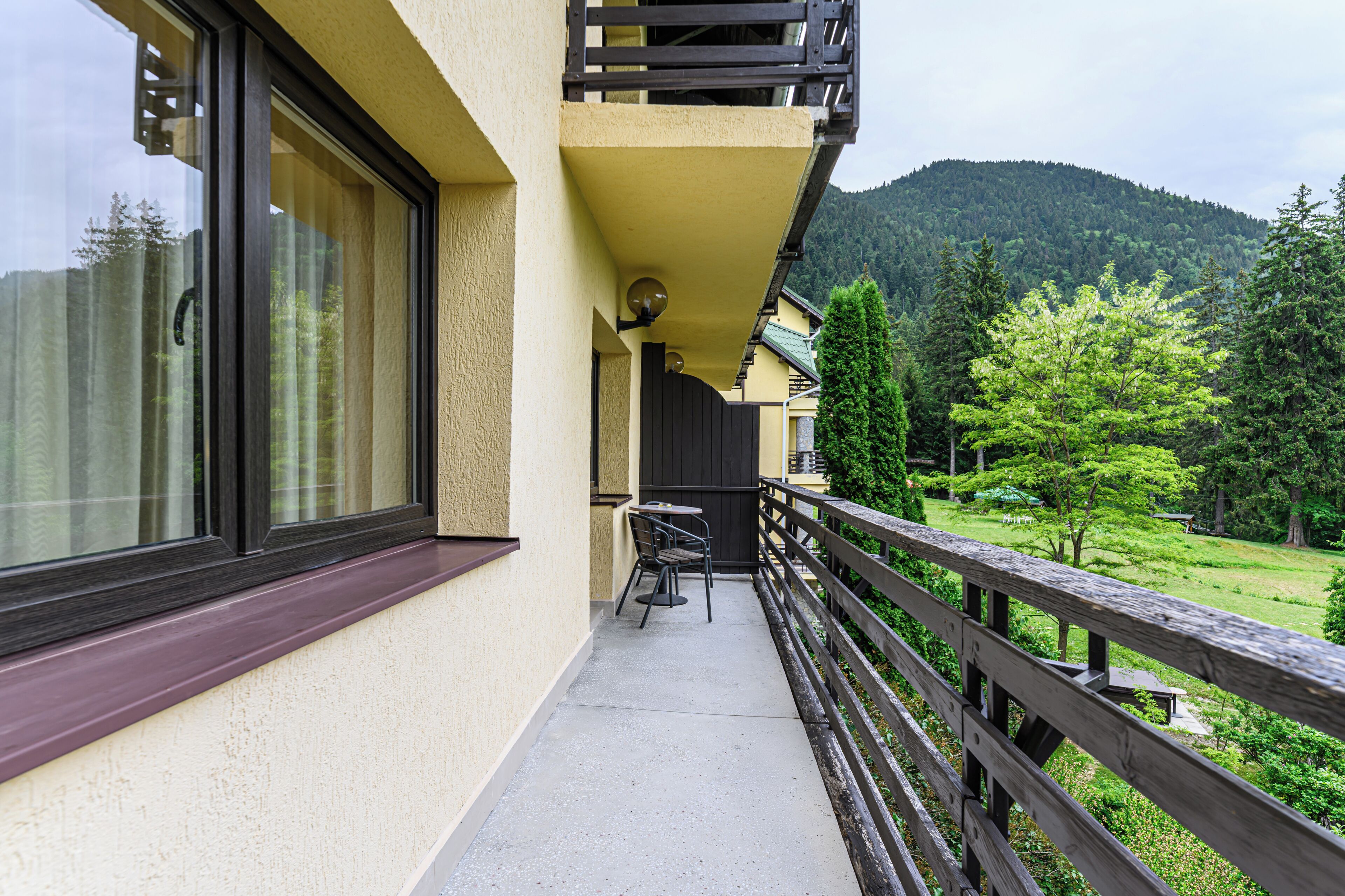 A balcony with a table and chairs, a wooden railing, and a view of a green landscape with trees and a hill in the background.