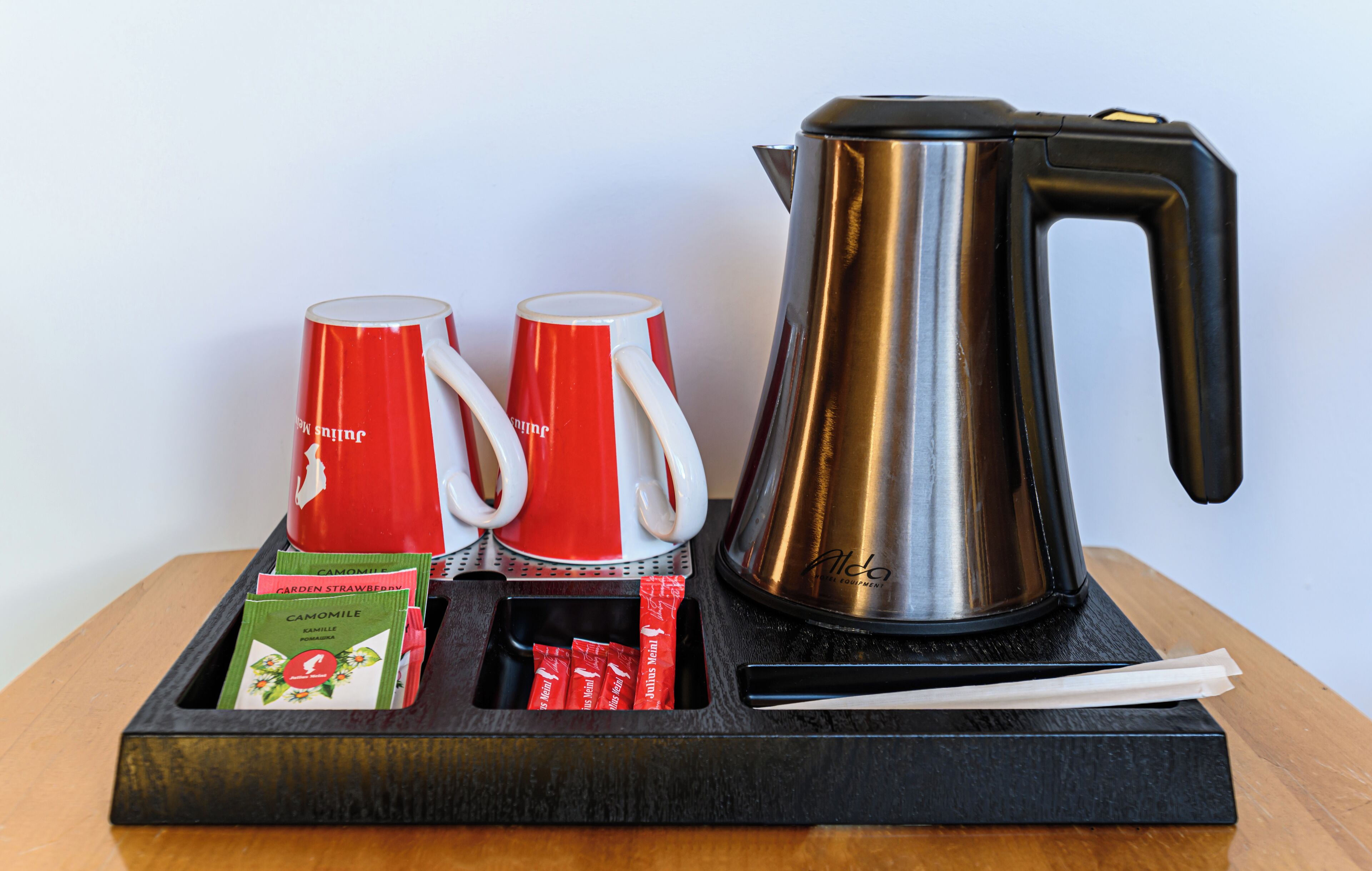 A coffee maker, two red cups, and a tray with tea bags and a spoon on a wooden table.