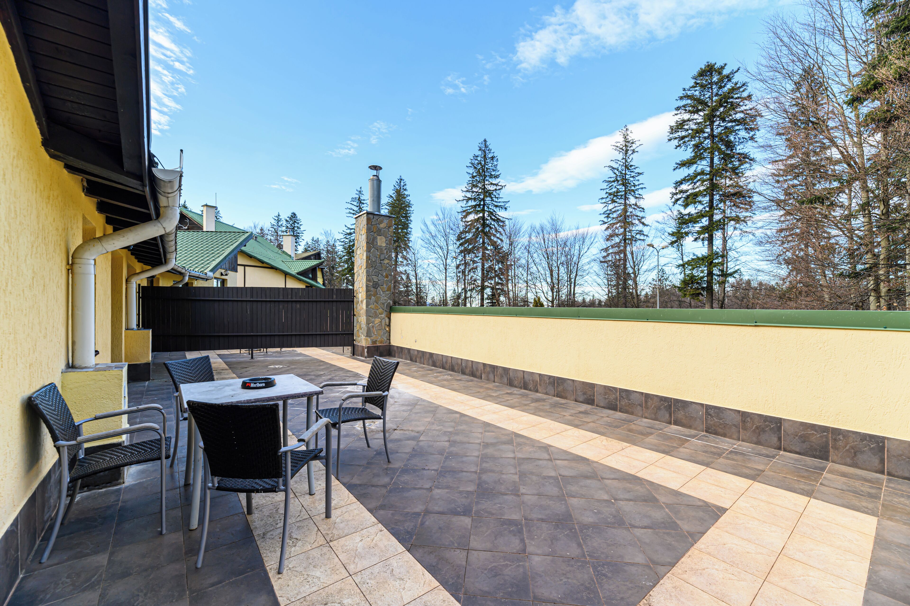 An outdoor patio with a table and chairs, a stone chimney, and a view of trees and a building in the background.