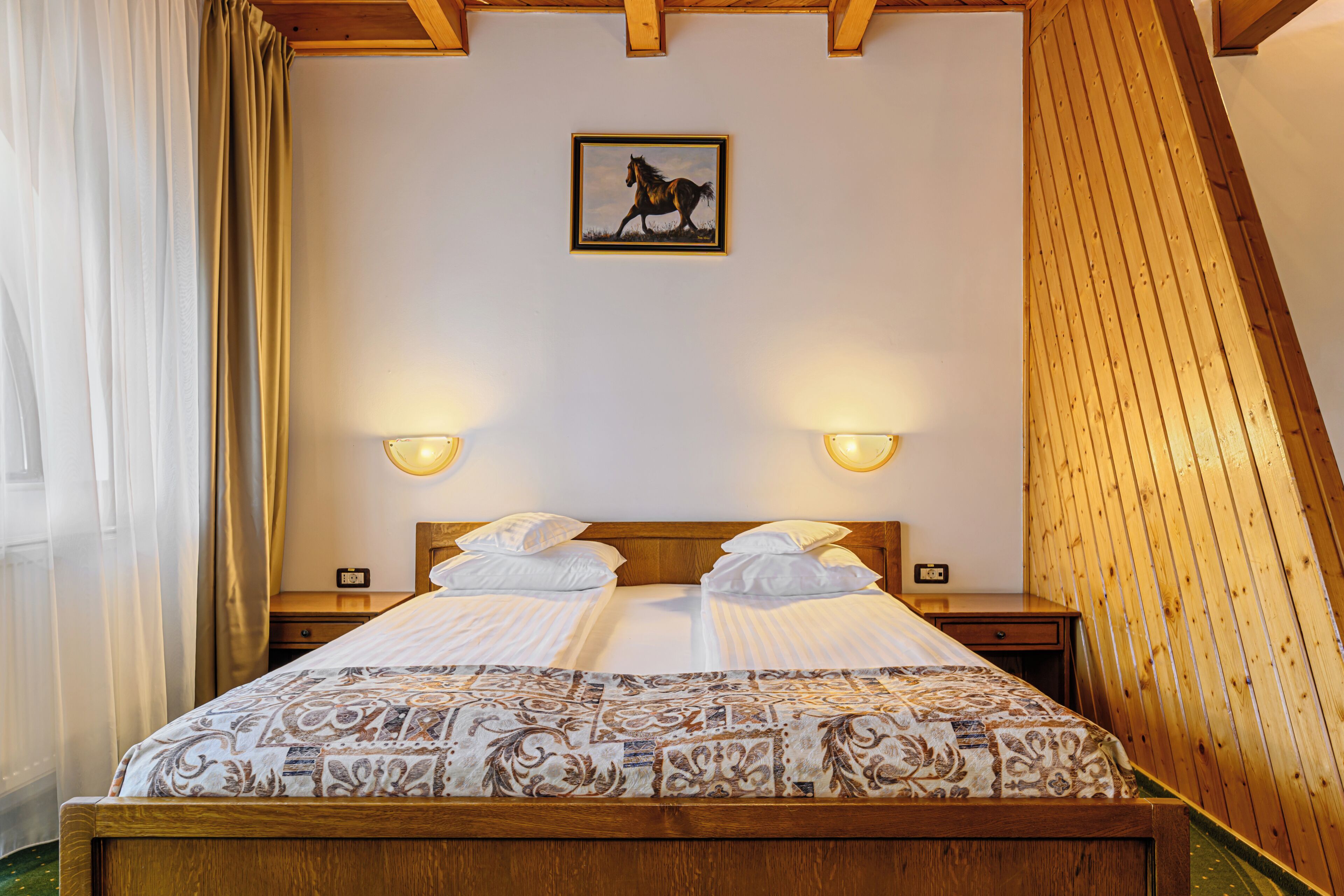 Una cama doble con una colcha estampada, dos mesitas de noche con lámparas, un cuadro enmarcado de un caballo en la pared y paneles de madera en el techo.