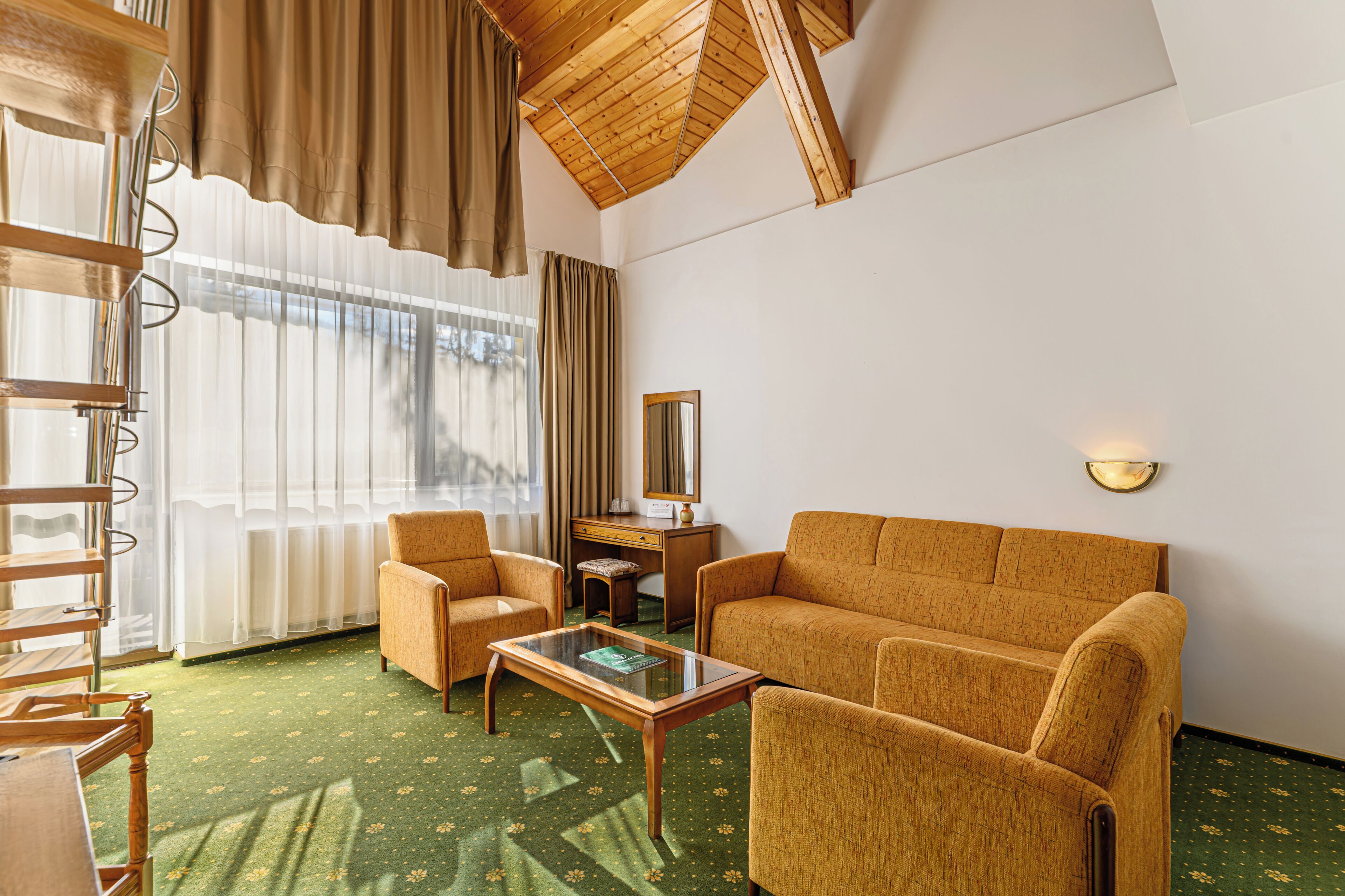 A room with a wooden ceiling, a green carpet, a sofa, two armchairs, a small table, a desk, a mirror, and a window with curtains.