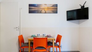 Un comedor con una mesa dispuesta para cuatro, sillas anaranjadas, un cuadro sobre la mesa y un televisor de pantalla plana en la pared.