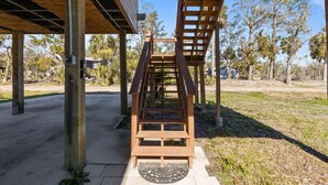 Un escalier extérieur en bois menant à une zone abritée, surmontée d’un toit en métal, et entouré d’arbres, avec un ciel dégagé.