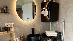 Ein modernes Badezimmer mit rundem Spiegel, Waschbecken in Schwarz, einer eingebauten Waschmaschine und einem Holzbodenregal.