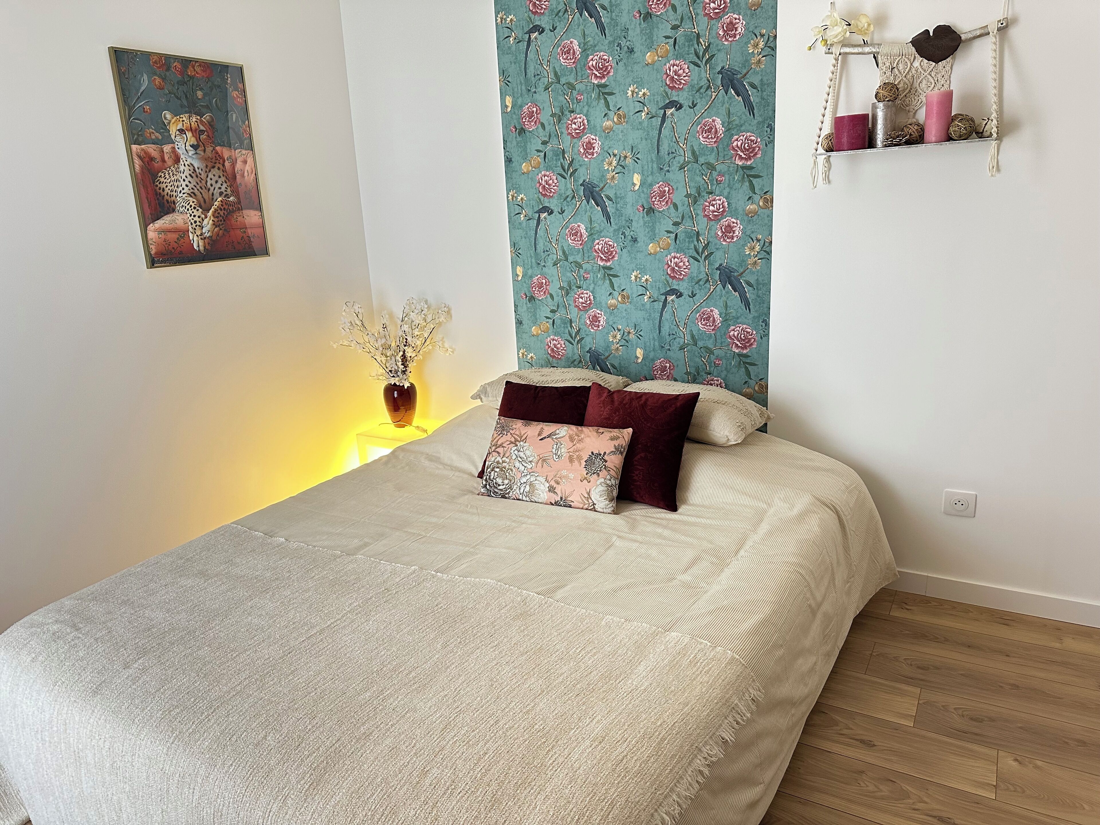 Een slaapkamer met een bed, een nachtkastje met een vaas, bloemenbehang en een schilderij aan de muur.