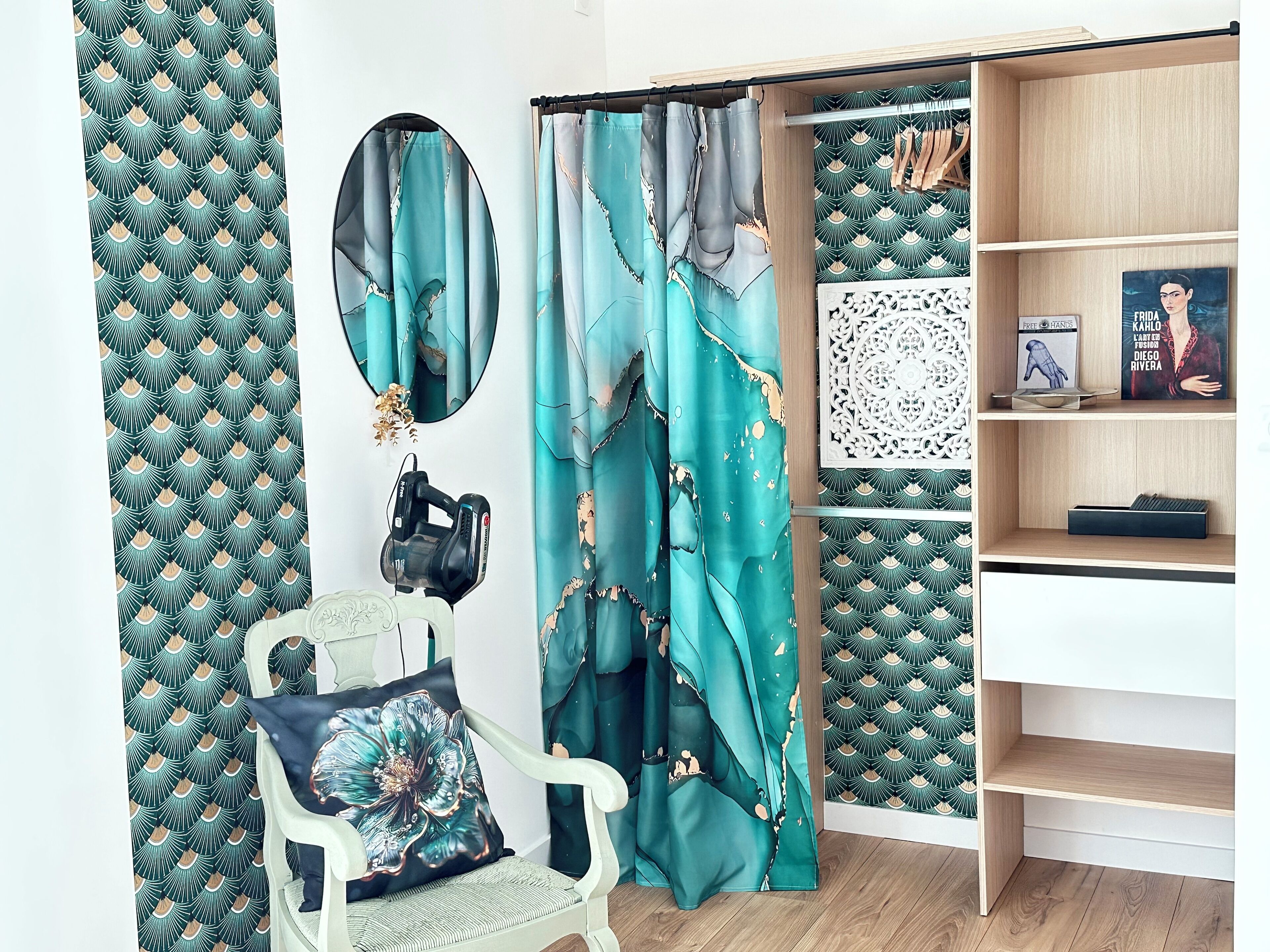 Een kamer met blauwgroene gordijnen met een patroon, een witte stoel met een bloemenkussen, een camera op een statief, en een boekenkast met boeken en decoratieve objecten.