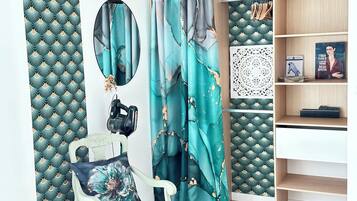 Een kamer met blauwgroene gordijnen met een patroon, een witte stoel met een bloemenkussen, een camera op een statief, en een boekenkast met boeken en decoratieve objecten.