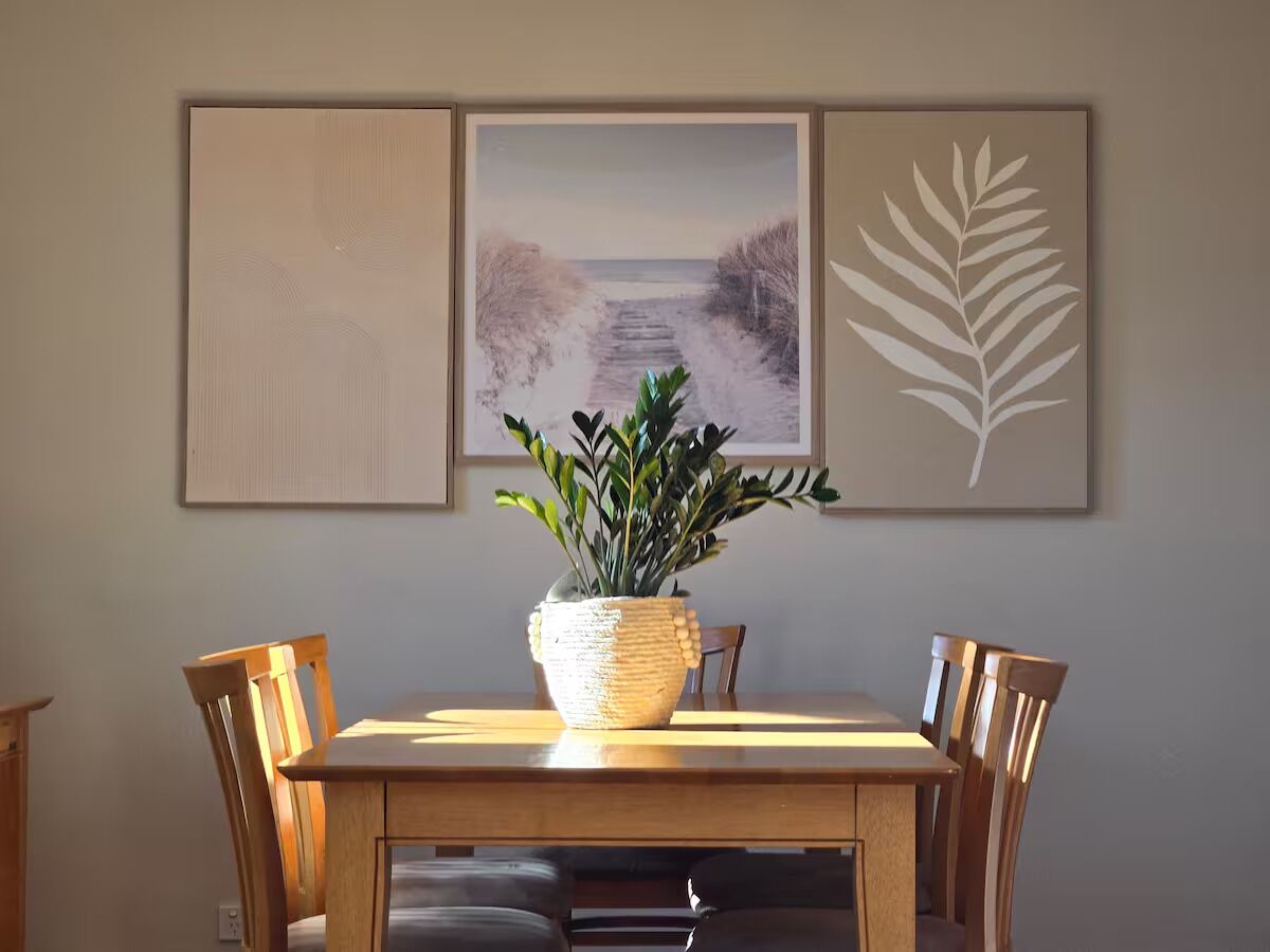 A dining area with a wooden table, chairs, a woven plant pot, and framed artwork on the wall.