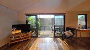 A spacious living area with a large sliding glass door leading to a patio, a wooden entertainment unit, a black leather sofa, and a desk with a lamp.