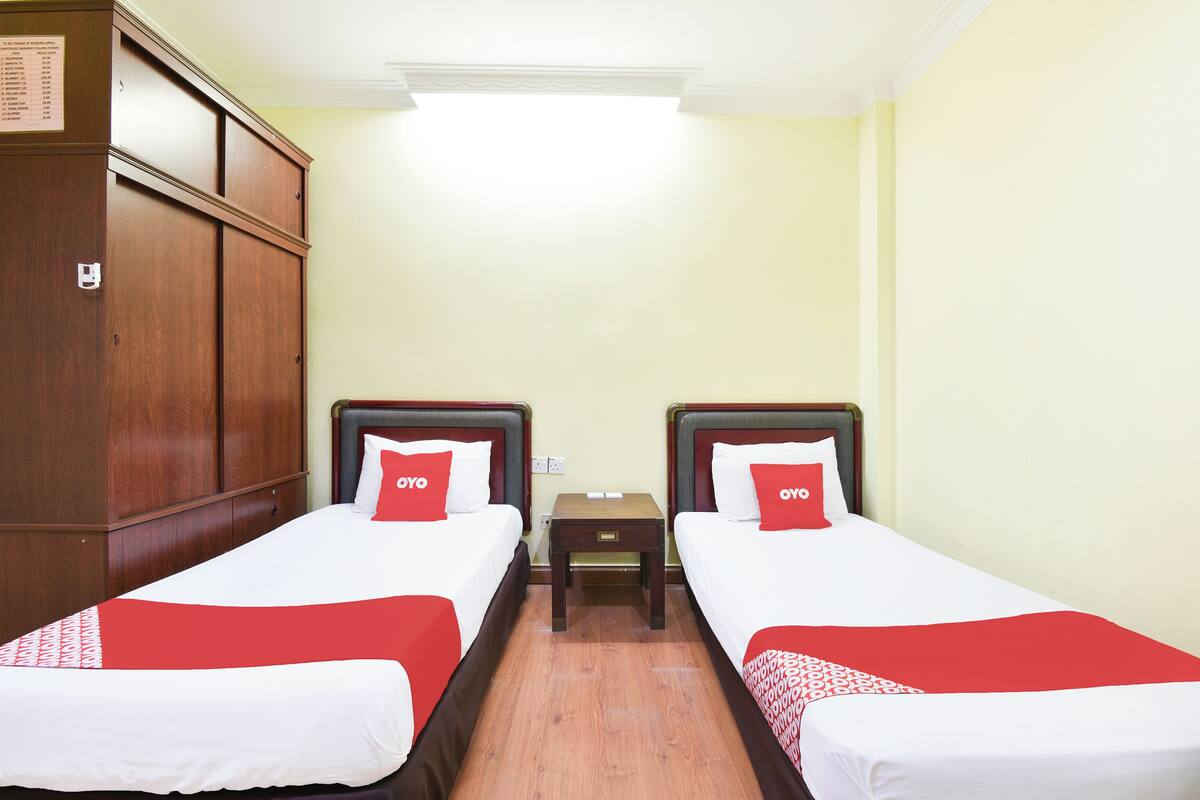 A hotel room with two beds, each with a red and white bedspread and matching pillows. There is a wooden wardrobe on the left and a small wooden nightstand between the beds.